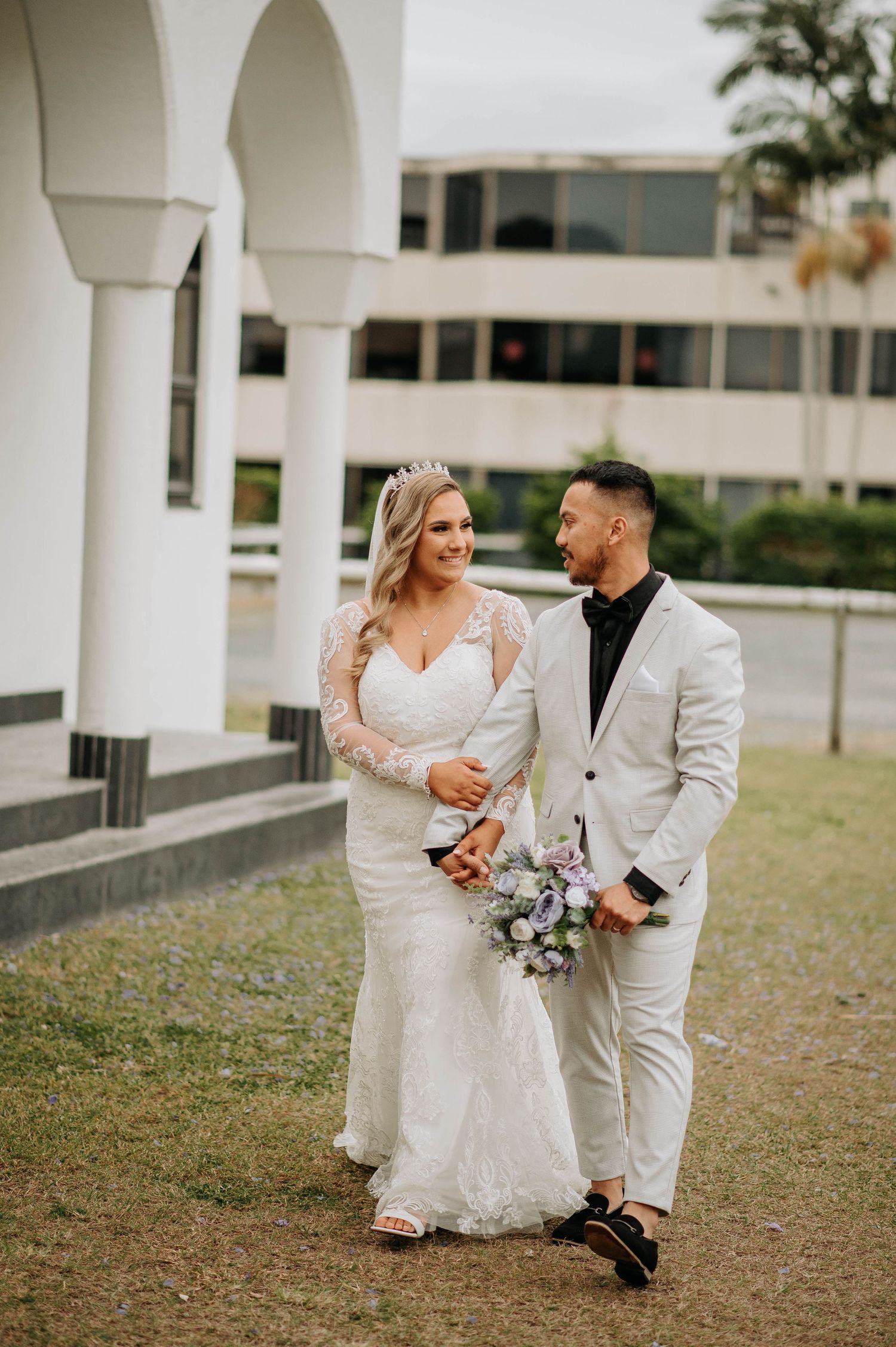 Newlywed couple walks together outdoors with white arched columns in background during wedding photoshoot.