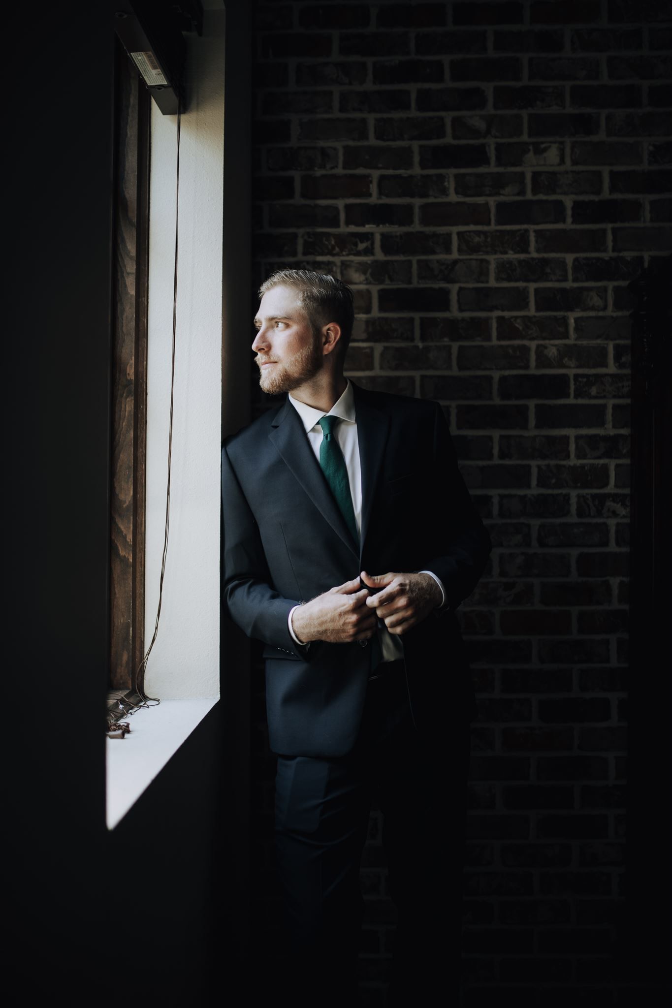 Professional figure in dark suit and green tie gazes out window against brick wall backdrop.
