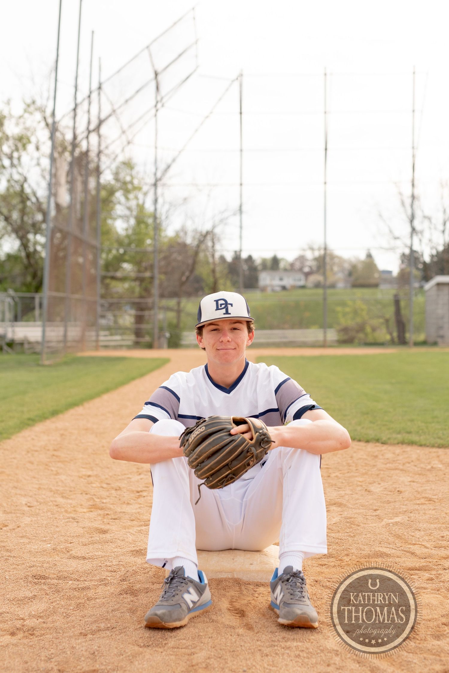 Baseball player sits on dirt field holding glove in white uniform with navy stripes.