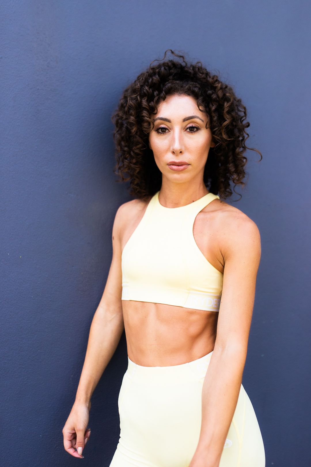 Athletic model in cream colored sports bra and shorts poses against blue wall background.