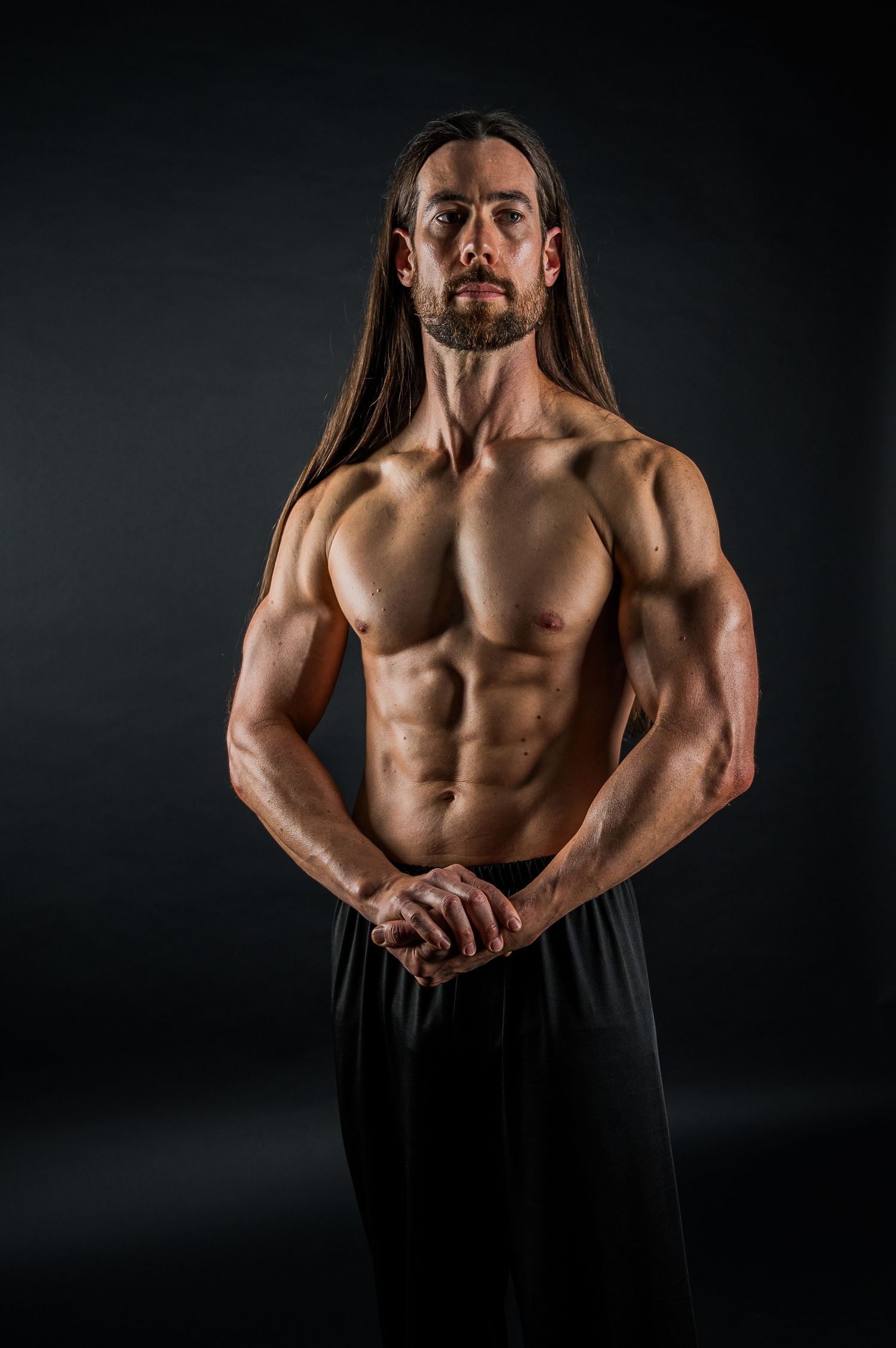 Fitness model with long hair displaying muscular physique against dark background.
