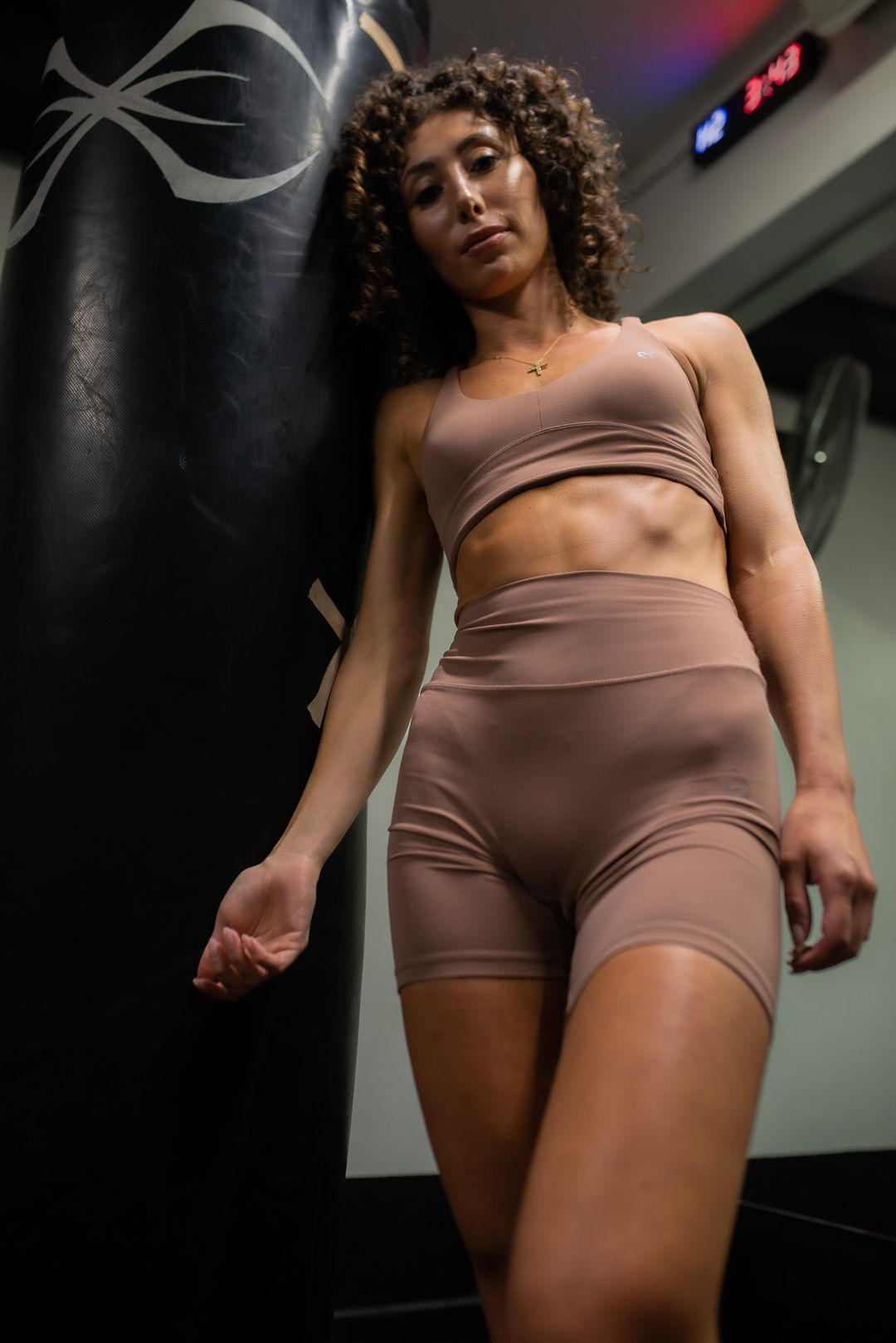Fitness model wearing brown workout set poses in dark gym setting with punching bag.