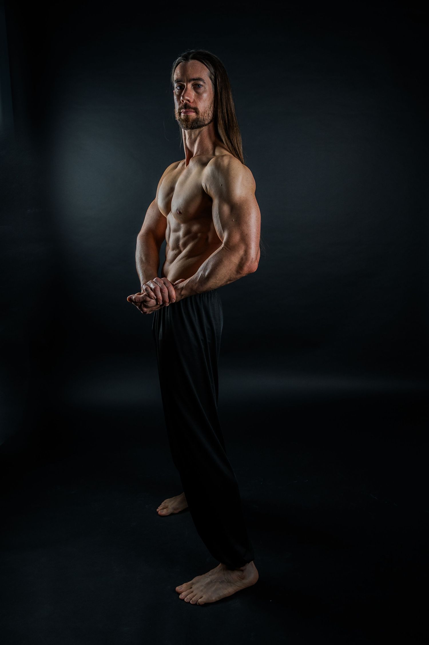 Muscular person with long hair poses shirtless against dark background in dramatic lighting.