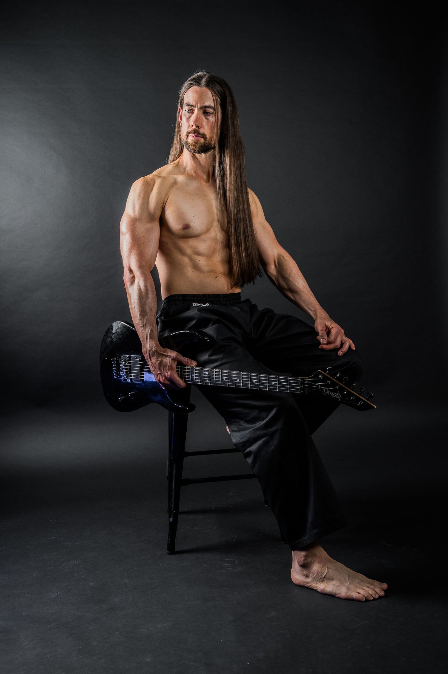 Muscular musician with long hair poses shirtless on chair with electric guitar against dark background.
