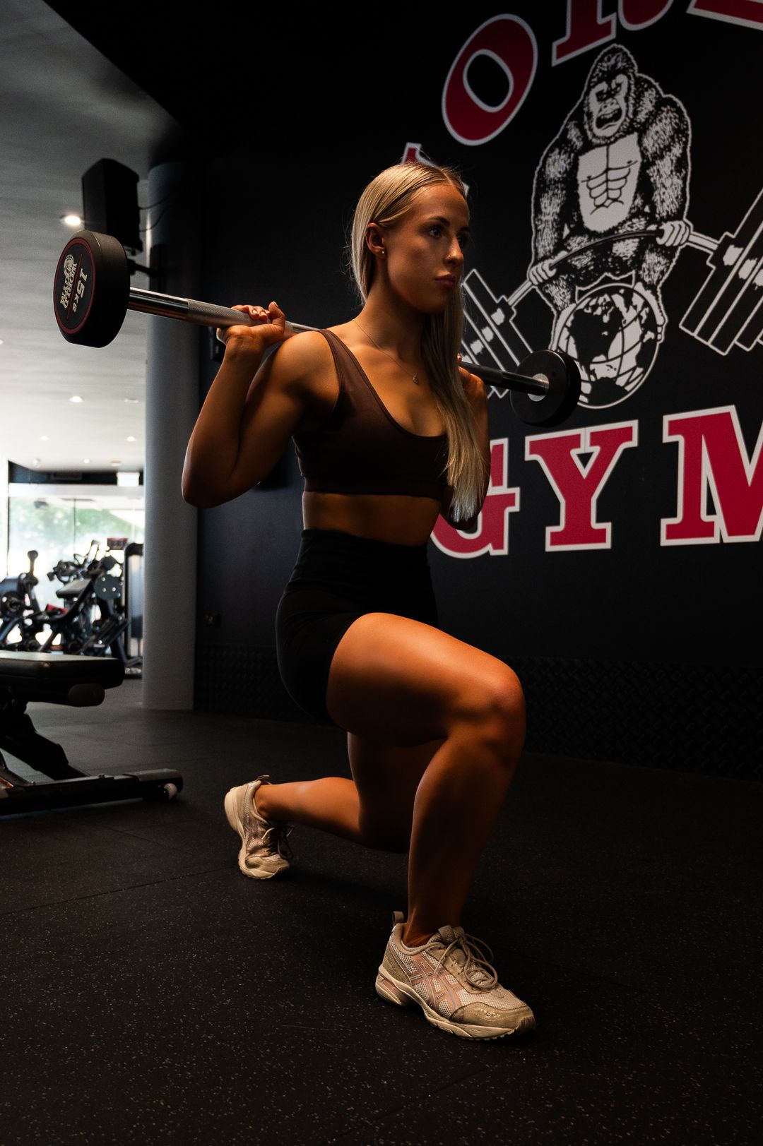 Fitness workout with barbell lunges against black gym wall with logo.