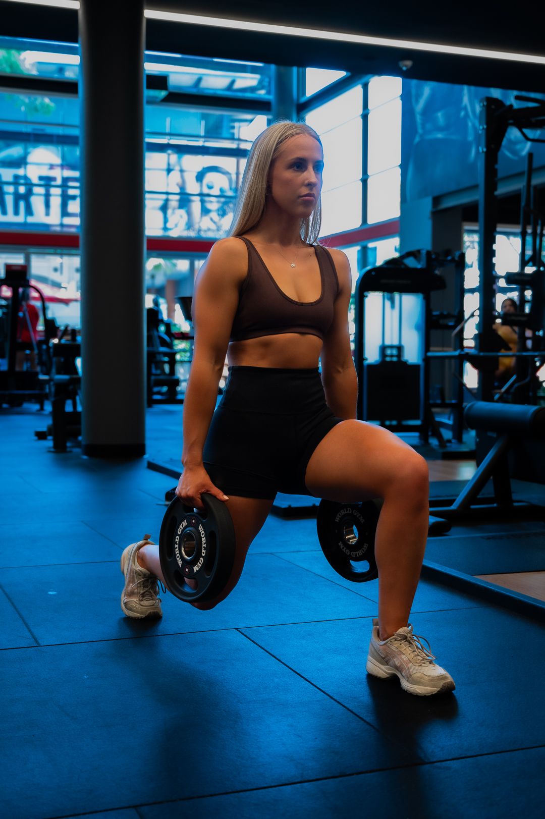 Woman performing dumbbell lunges in a blue-lit modern gym setting.