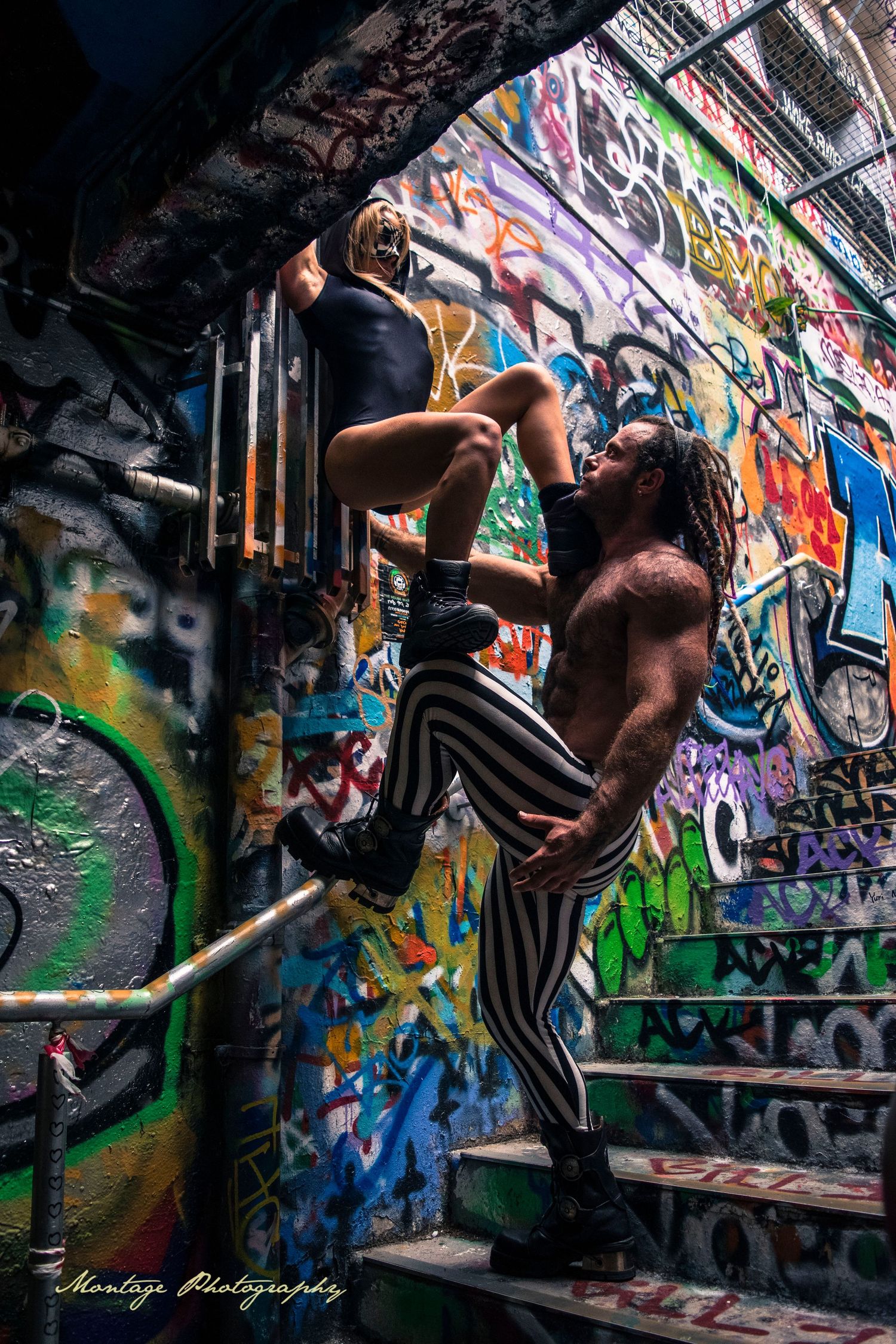 Dancers execute dramatic lift pose against backdrop of graffiti-covered stairwell.