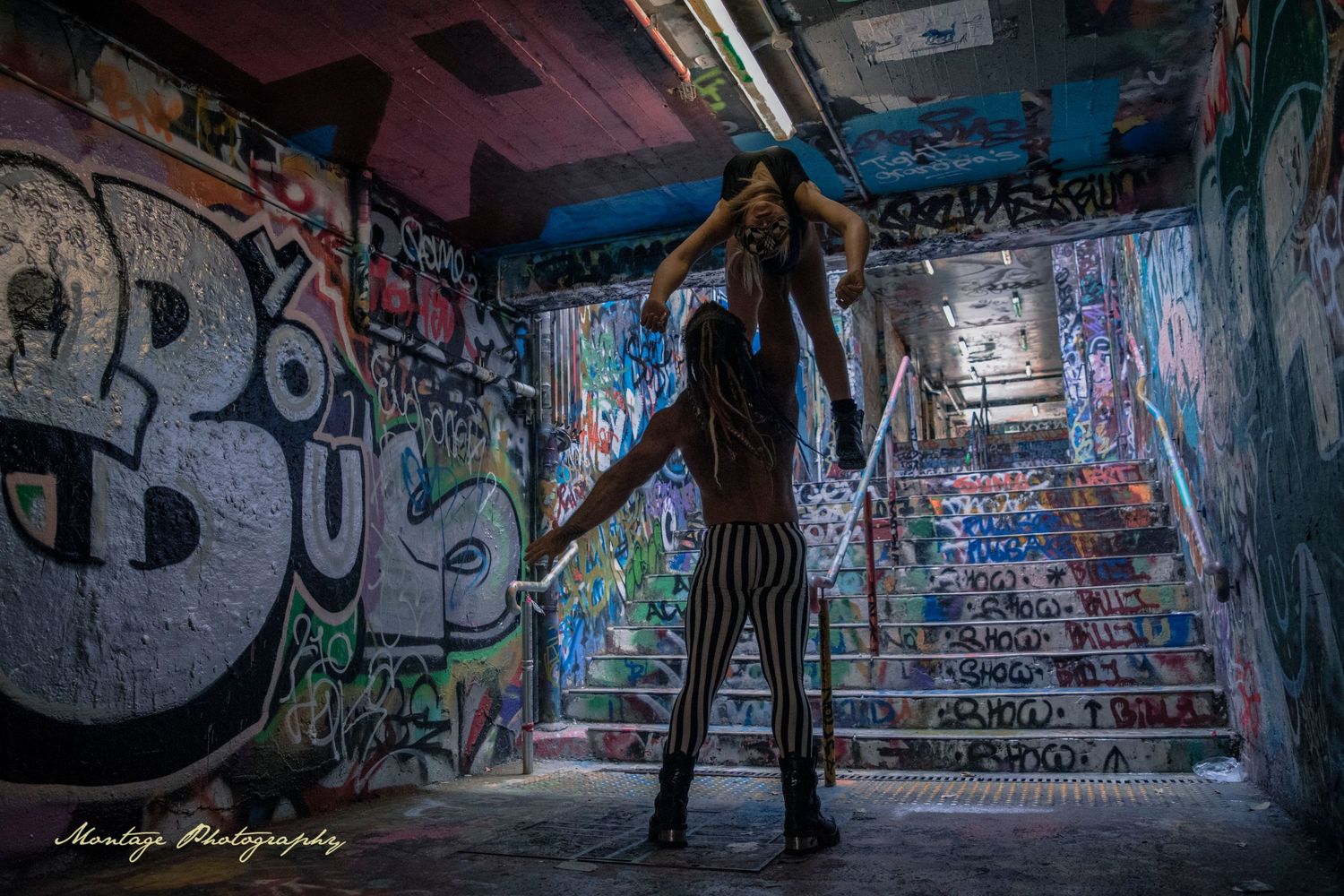 Acro figures create dramatic pose in graffiti-covered tunnel with converging lines.
