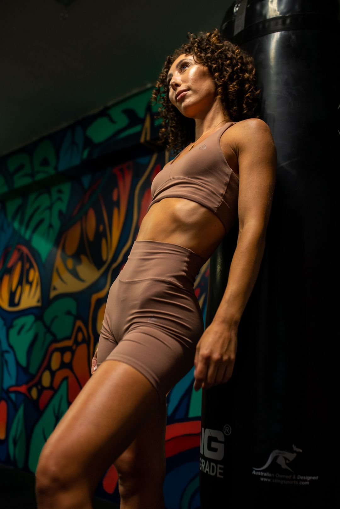 Athletic figure in brown two-piece workout set poses against colorful graffiti wall in gym setting.