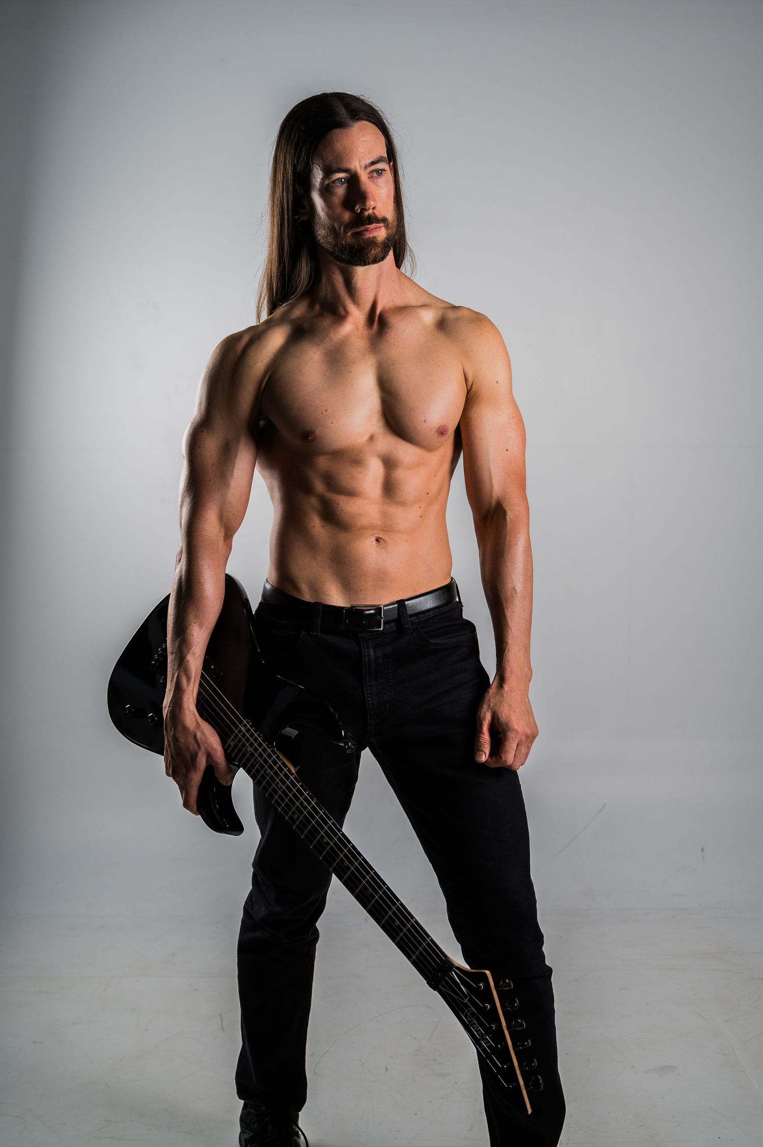 Athletic guitarist with long dark hair holds electric guitar while wearing black pants in studio setting.