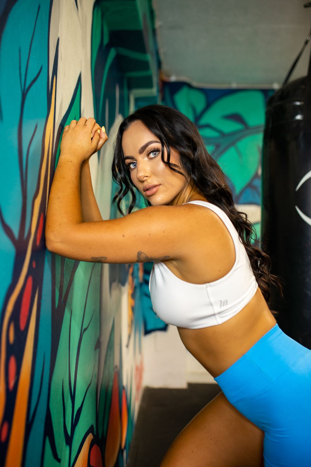 Athletic figure in workout attire posing against colorful mural wall in gym setting.
