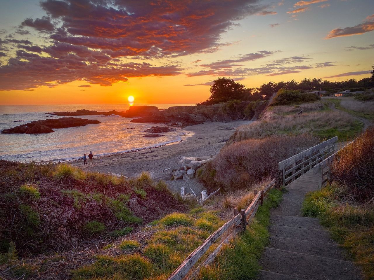 Dramatic sunset over coastal path with wooden bridge and vibrant orange sky.