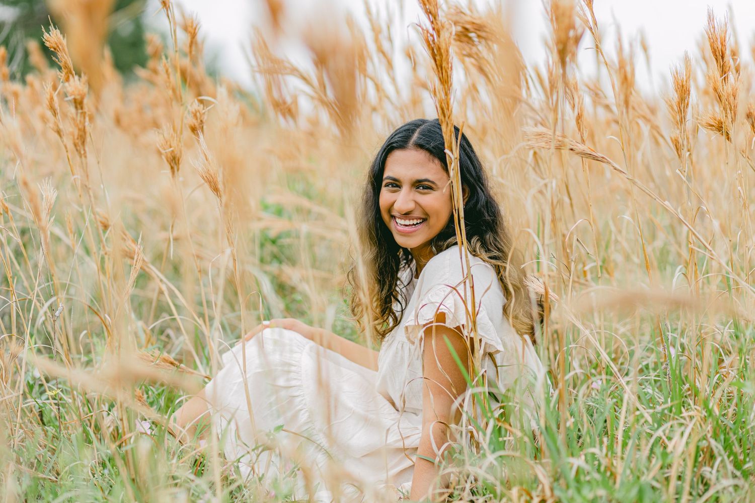 A highschool senior smiles at the camera sitting in a field of tall grass.