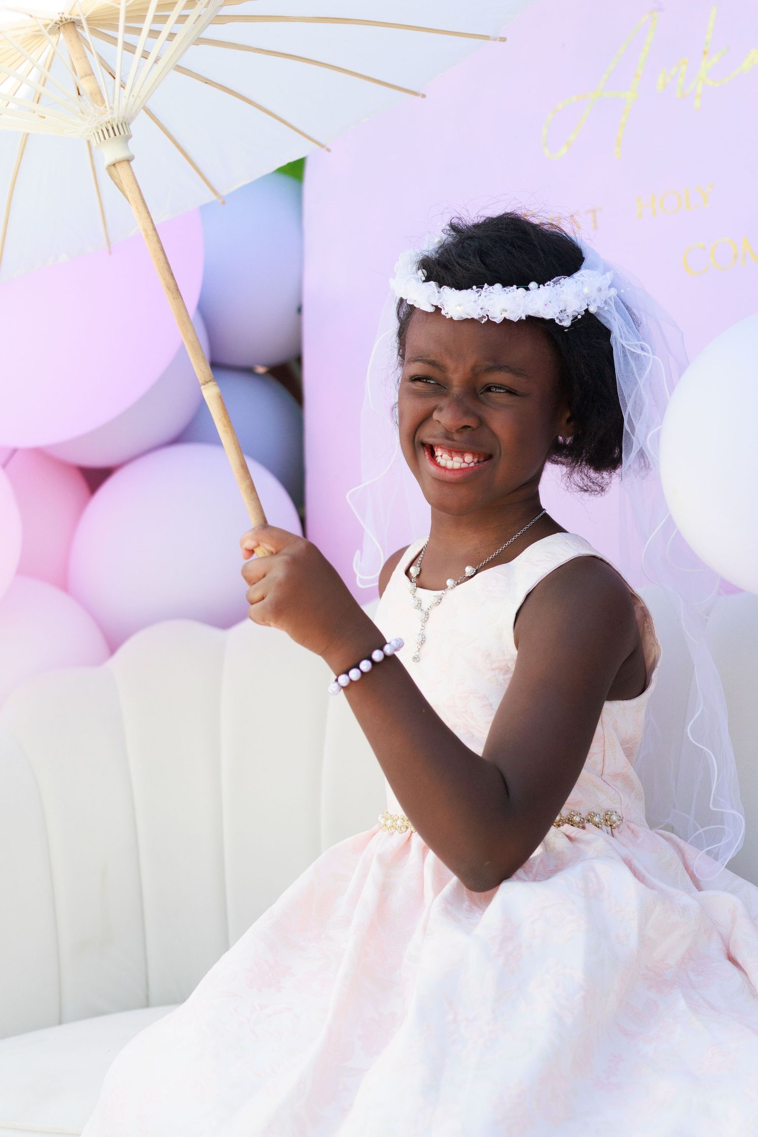 A child in a white dress and veil holding a decorative parasol poses against a pastel balloon backdrop.