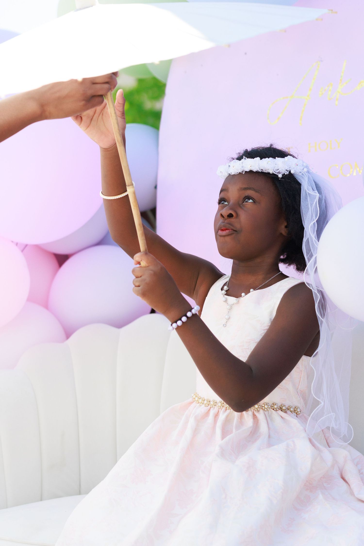 A young girl in first communion attire holds a delicate parasol against a backdrop of pastel balloons.
