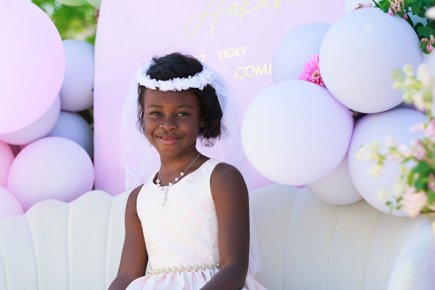 A child wearing a white dress and headpiece smiles for a portrait with pastel balloons in the background.