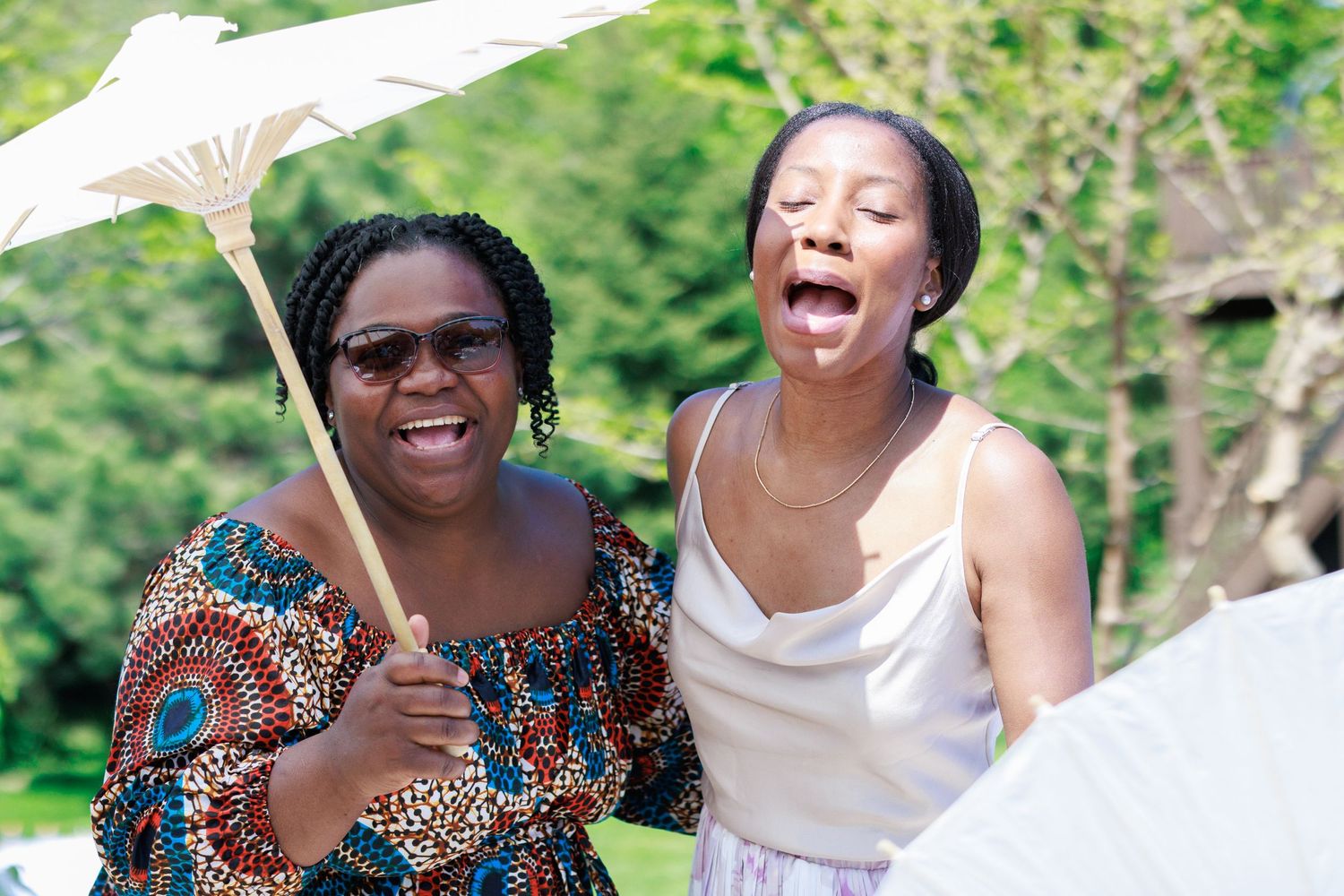 Two friends sharing joyful laughter while holding a white parasol at an outdoor event.