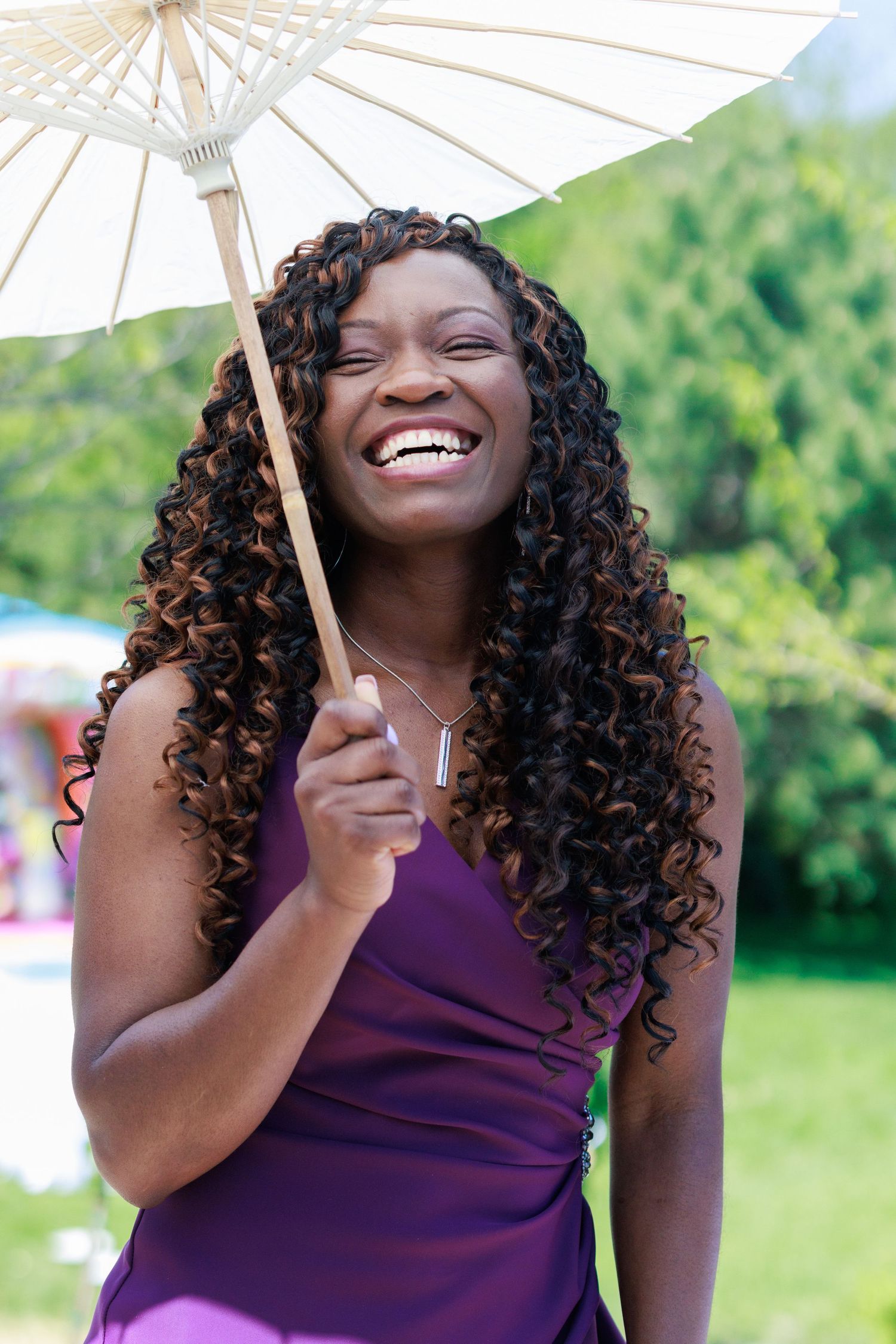 Woman in purple dress smiling while holding white parasol at outdoor celebration.