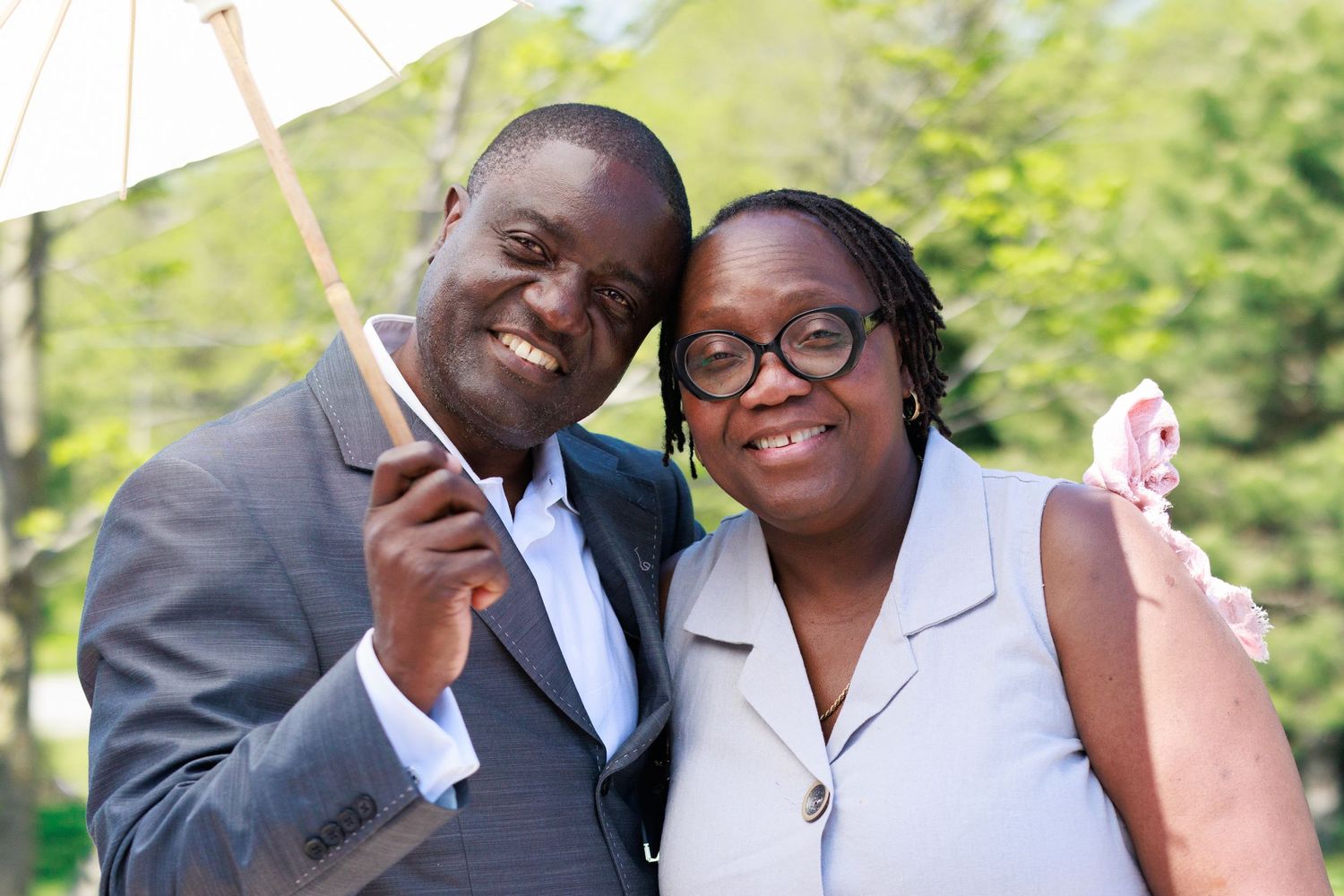 Couple in formal attire sharing a white parasol at outdoor gathering.