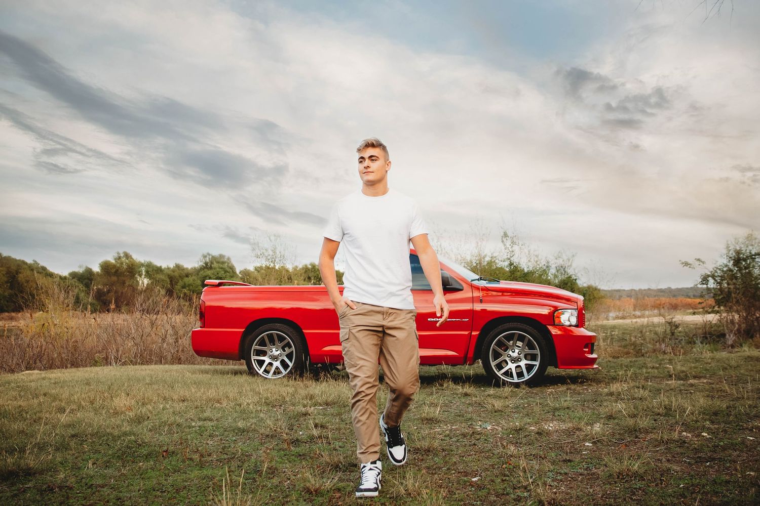 Senior boy stands beside red vintage pickup truck in grassy field at sunset.