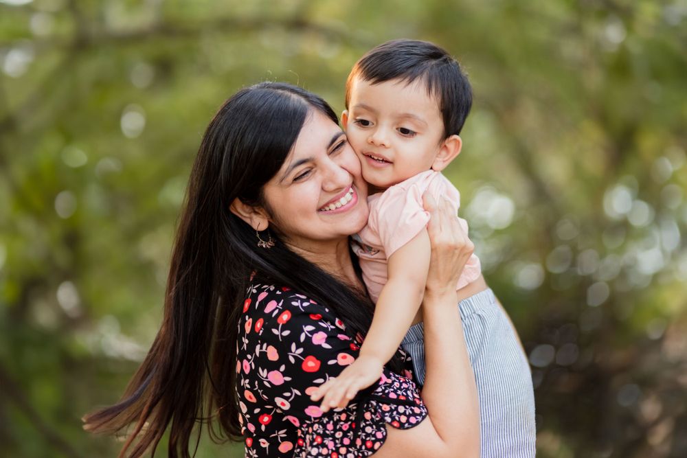 A person in a floral dress embraces a toddler outdoors against a soft green bokeh background.