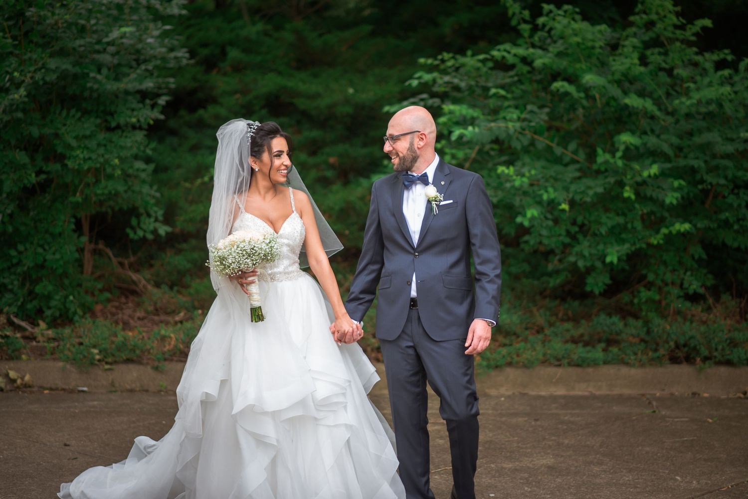 Newlywed couple walks hand in hand through a lush green wooded outdoor setting.