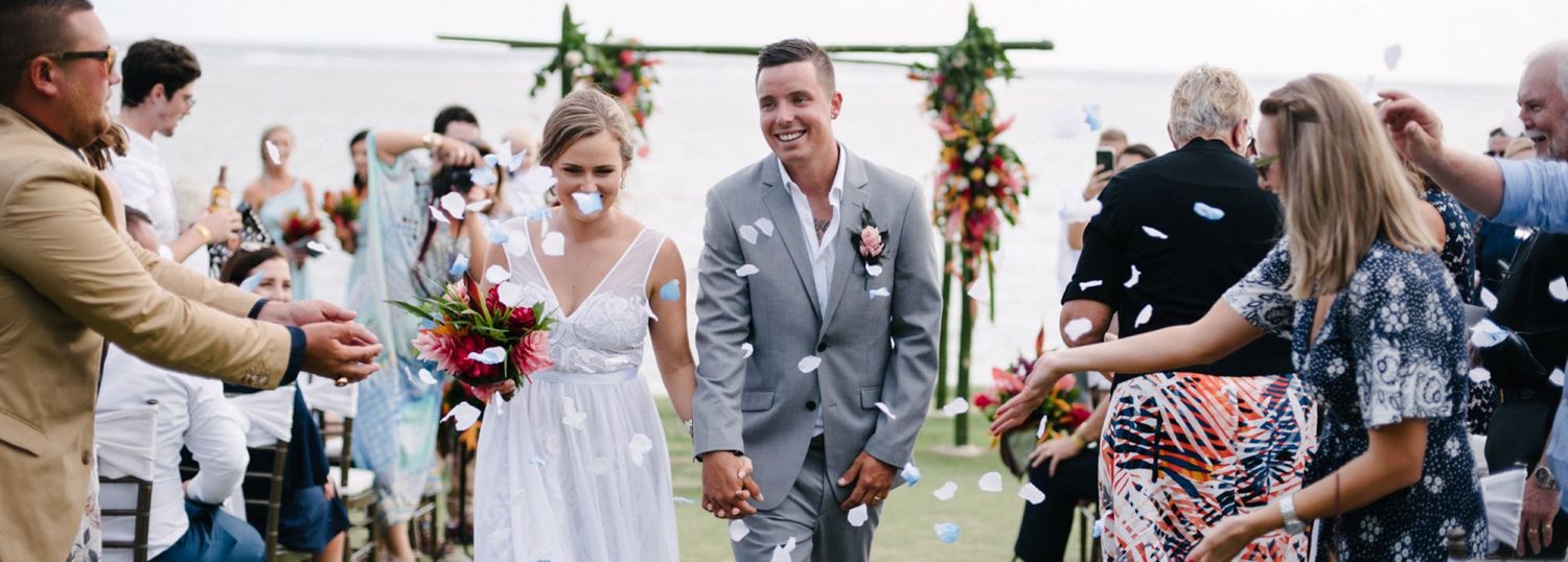 Joyful wedding guests throw flower petals as a couple exits their outdoor ceremony under hanging greenery.