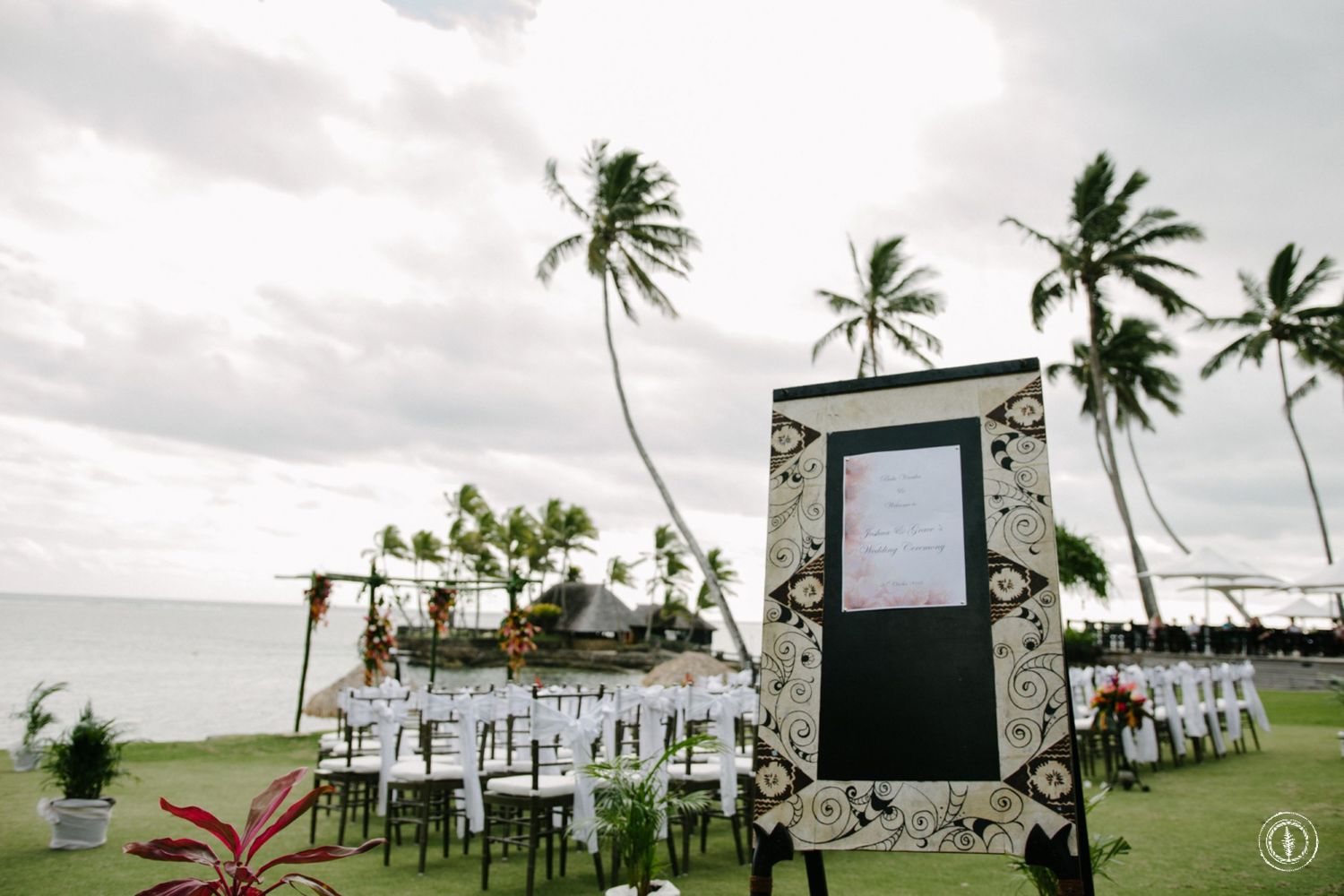 Tropical beach wedding setup with white chairs and stone-framed sign surrounded by swaying palm trees.