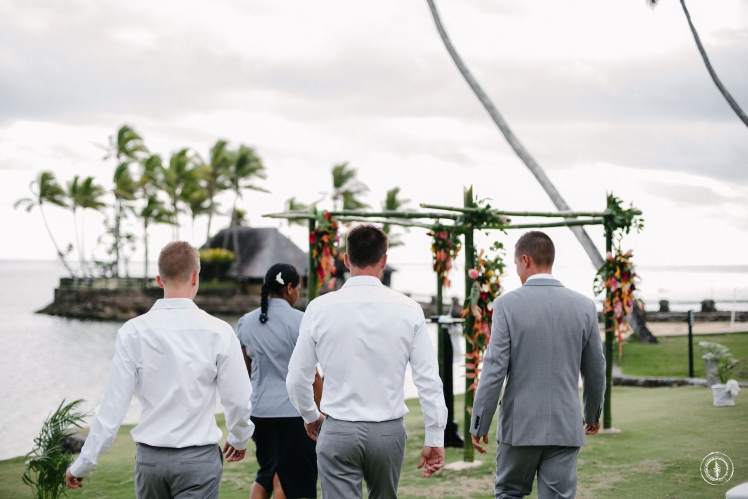 Small group walking towards a flower-decorated wedding arch with ocean and palm trees in background.