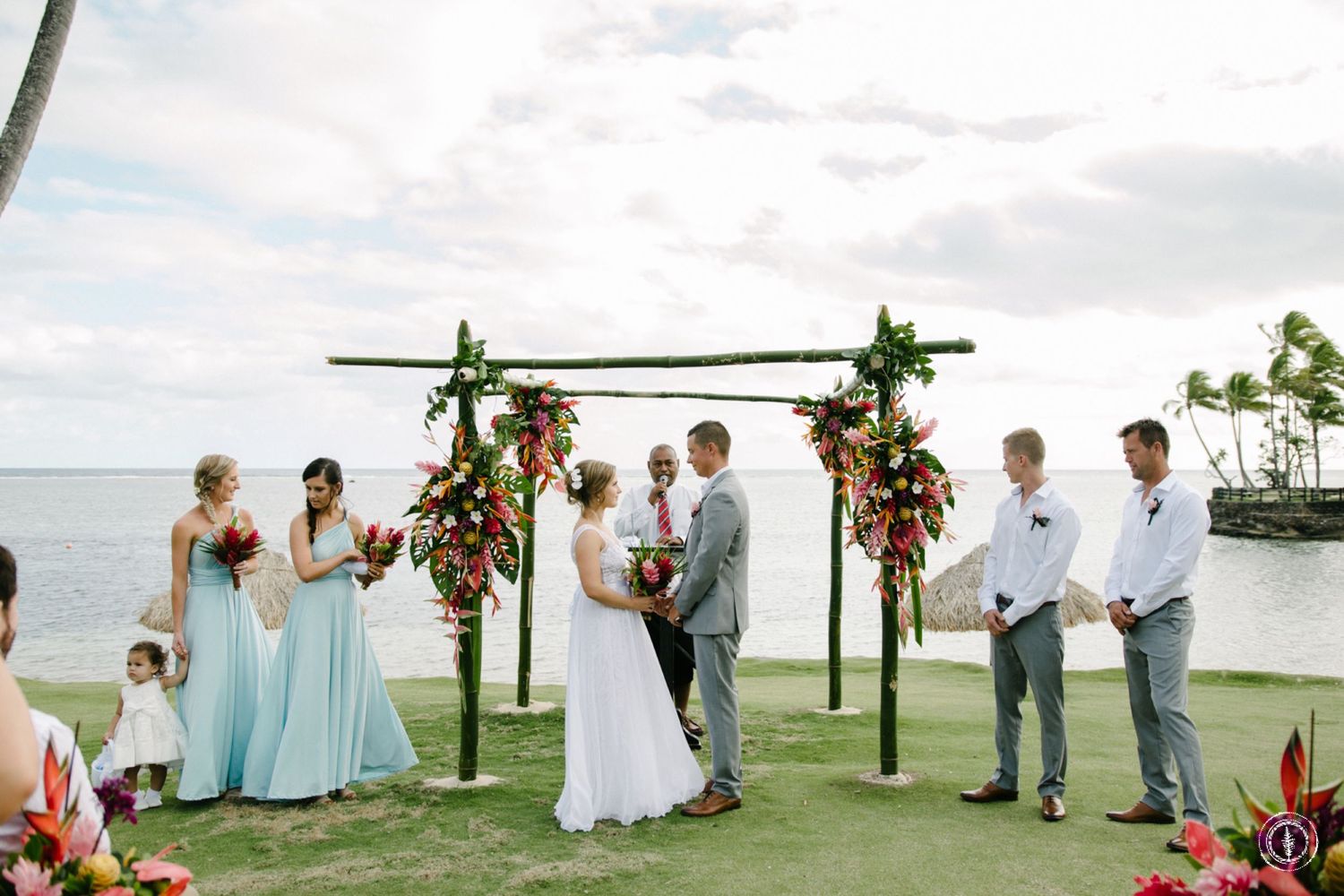 Outdoor beach wedding ceremony under a wooden arch decorated with tropical flowers.