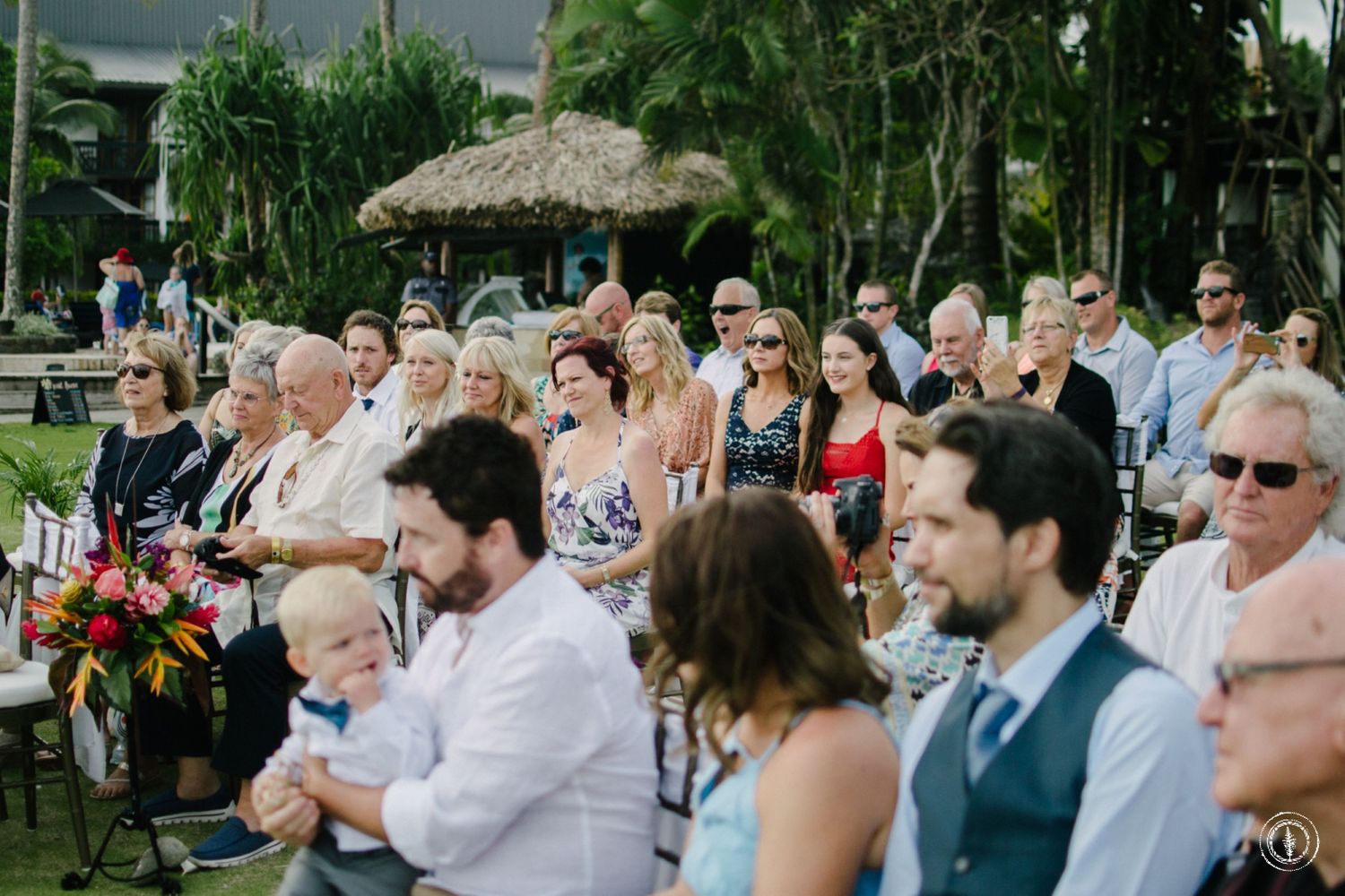 Wedding guests seated on lawn chairs at an outdoor tropical ceremony.