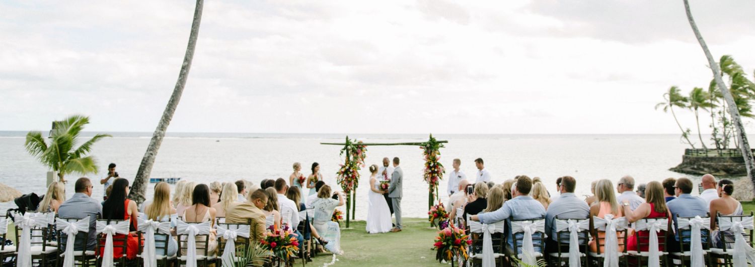 Panoramic view of a beachside wedding ceremony with palm trees and white chairs.