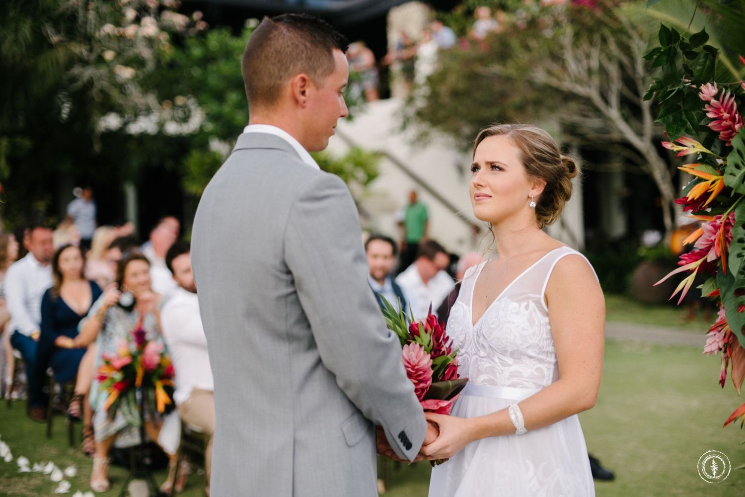 A couple exchanges vows during an outdoor garden wedding ceremony surrounded by tropical flowers and guests.
