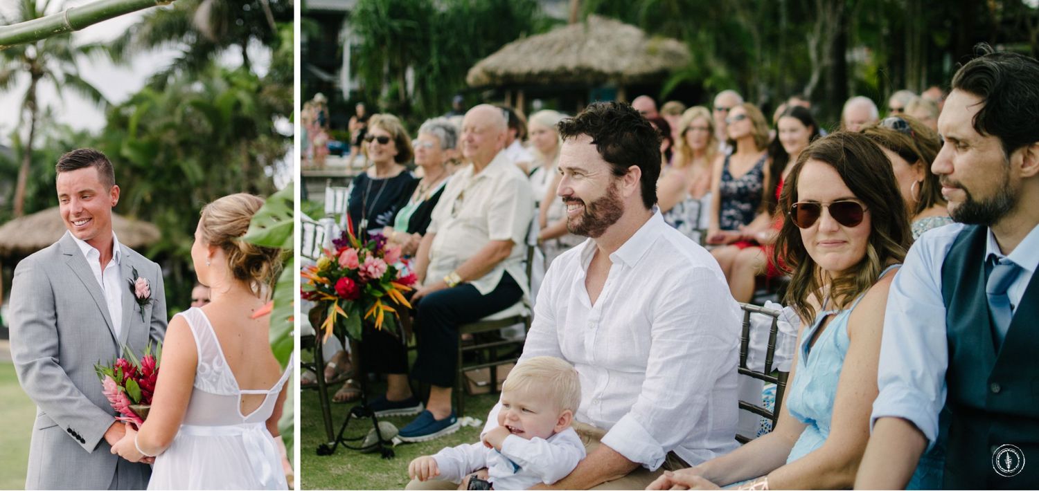 Wedding guests seated in white chairs watch an outdoor ceremony under palm trees.