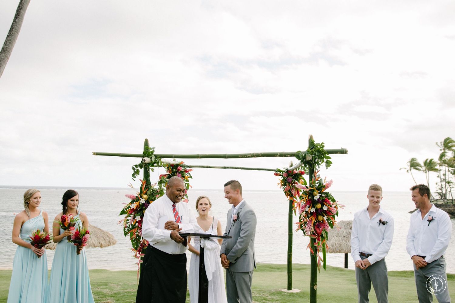 Wedding ceremony on a lawn overlooking the ocean with bamboo arch and tropical flowers.