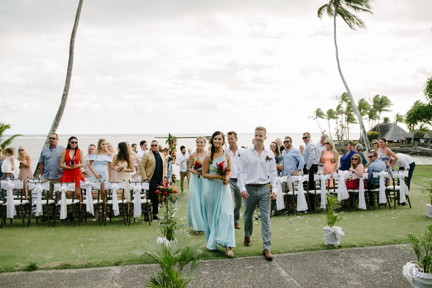 Wedding ceremony guests seated on white chairs along a palm-lined tropical oceanfront lawn.