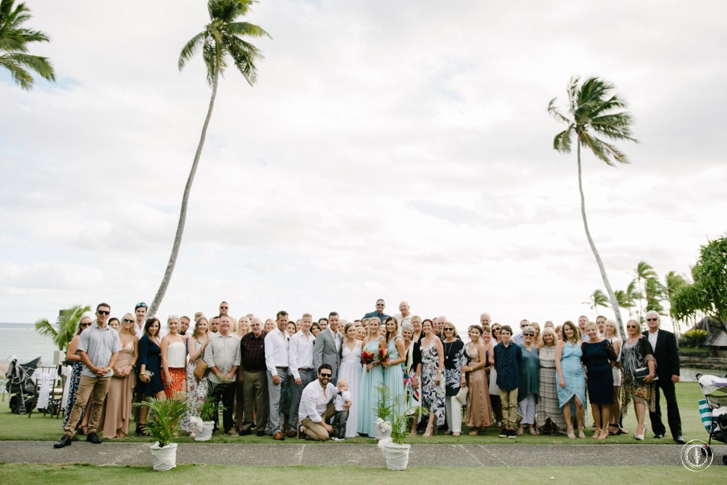 Large wedding group photo with guests on tropical lawn with swaying palm trees against cloudy sky.