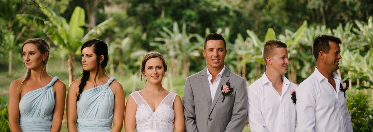 Wedding party in light blue and gray attire standing together in tropical garden setting.