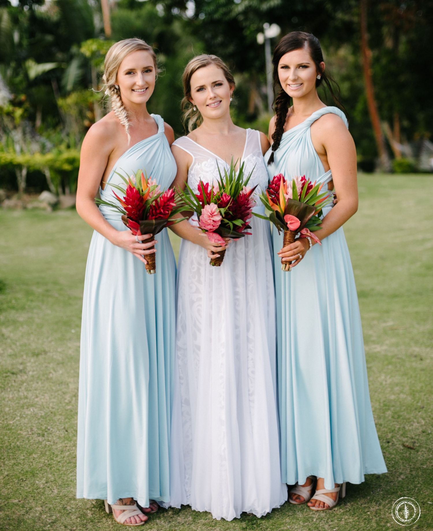Three bridesmaids in light blue gowns hold colorful tropical bouquets while posing on a grassy lawn.