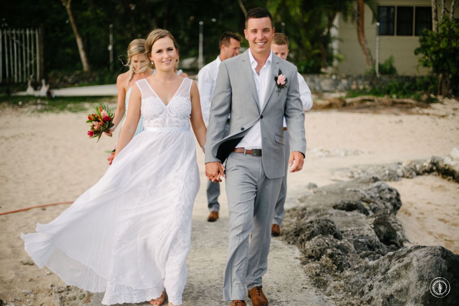 A couple walks along a tropical beach, with the bride's white dress flowing in the breeze.