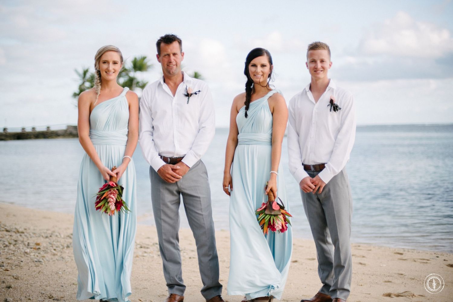 A wedding party poses on a beach with light blue dresses and grey suits against an ocean backdrop.
