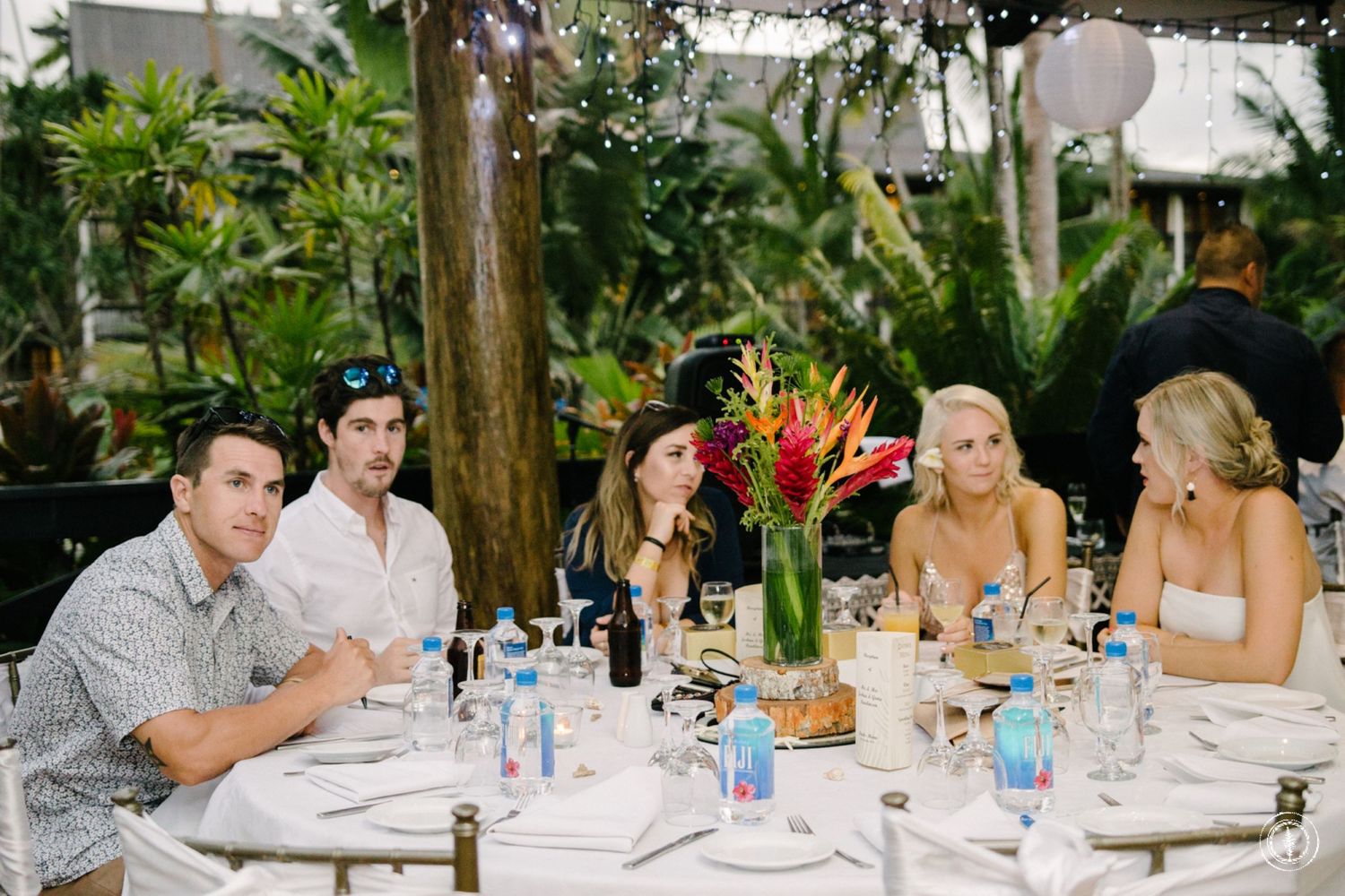 Outdoor tropical wedding reception with guests dining at decorated tables with floral centerpieces.