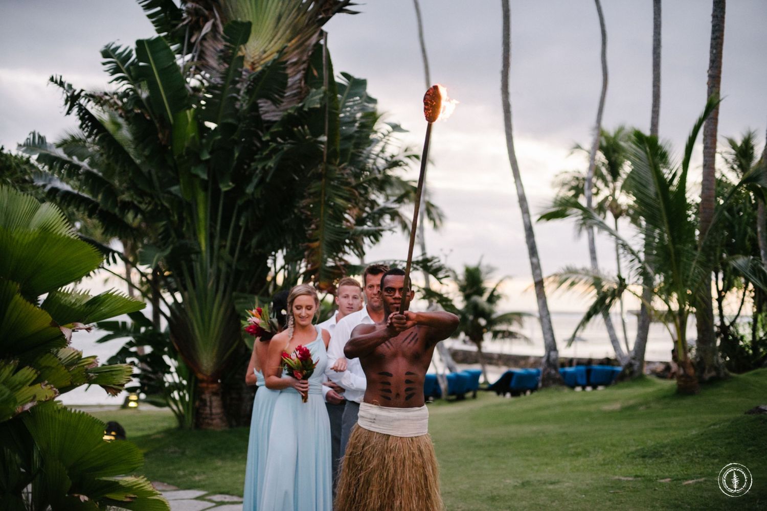 Traditional Hawaiian fire dance performance on a lush beachfront lawn during sunset.