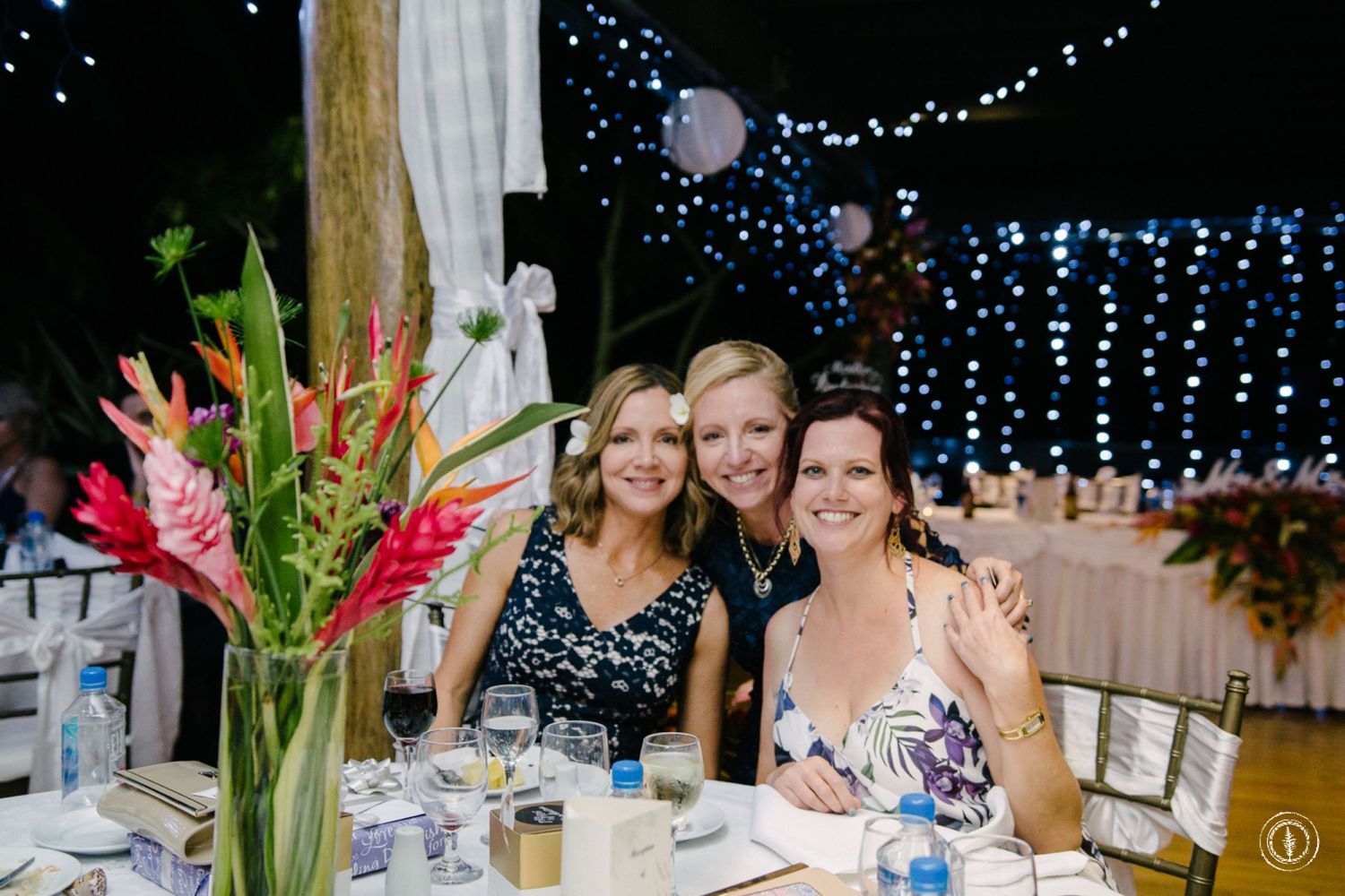 Wedding reception dinner under blue string lights with tropical floral centerpiece and guests at decorated tables.