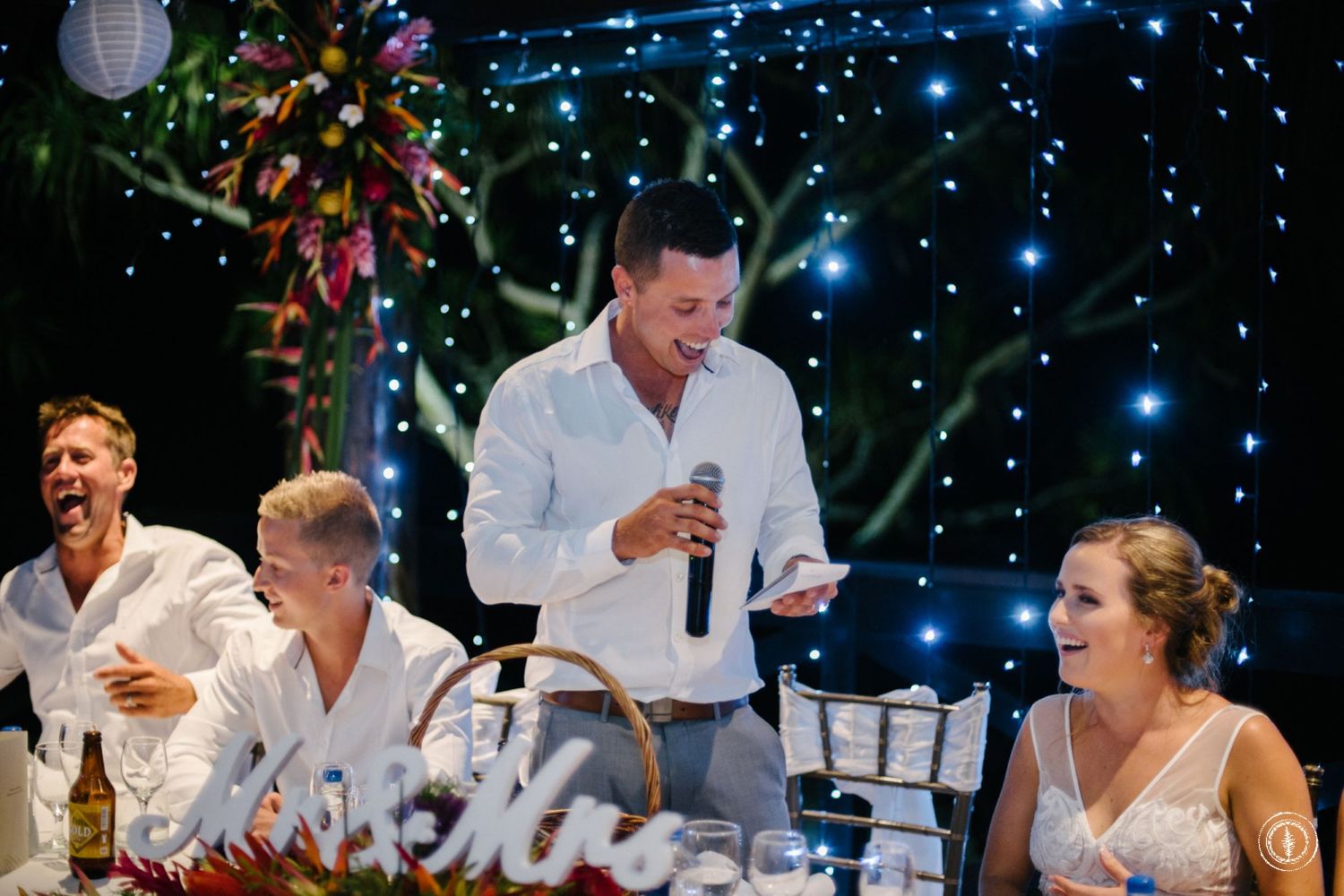 Wedding reception guests laughing during speeches under twinkling blue string lights.
