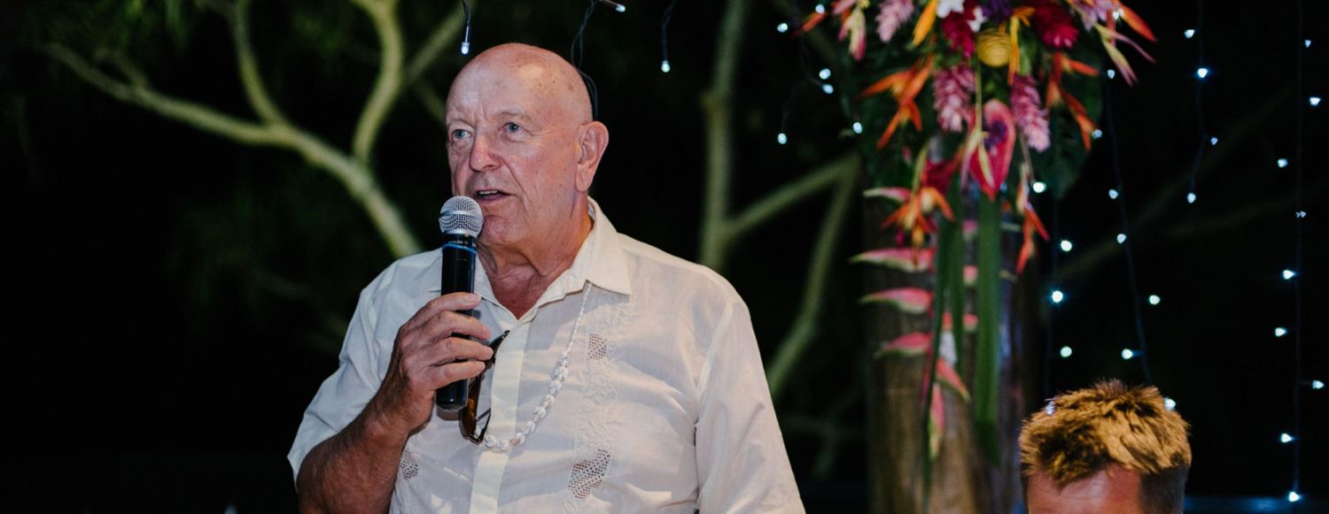 Speaker giving speech at outdoor wedding reception with tropical floral arrangements and string lights.