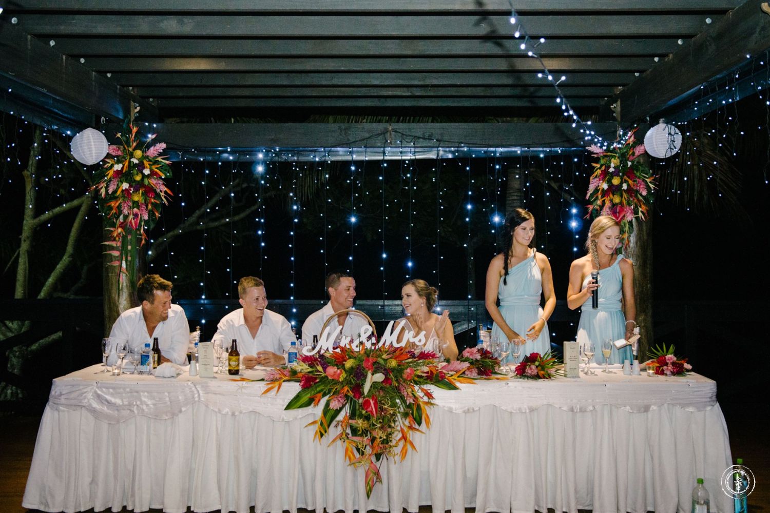 Wedding party seated at head table with tropical floral arrangements and string light backdrop.
