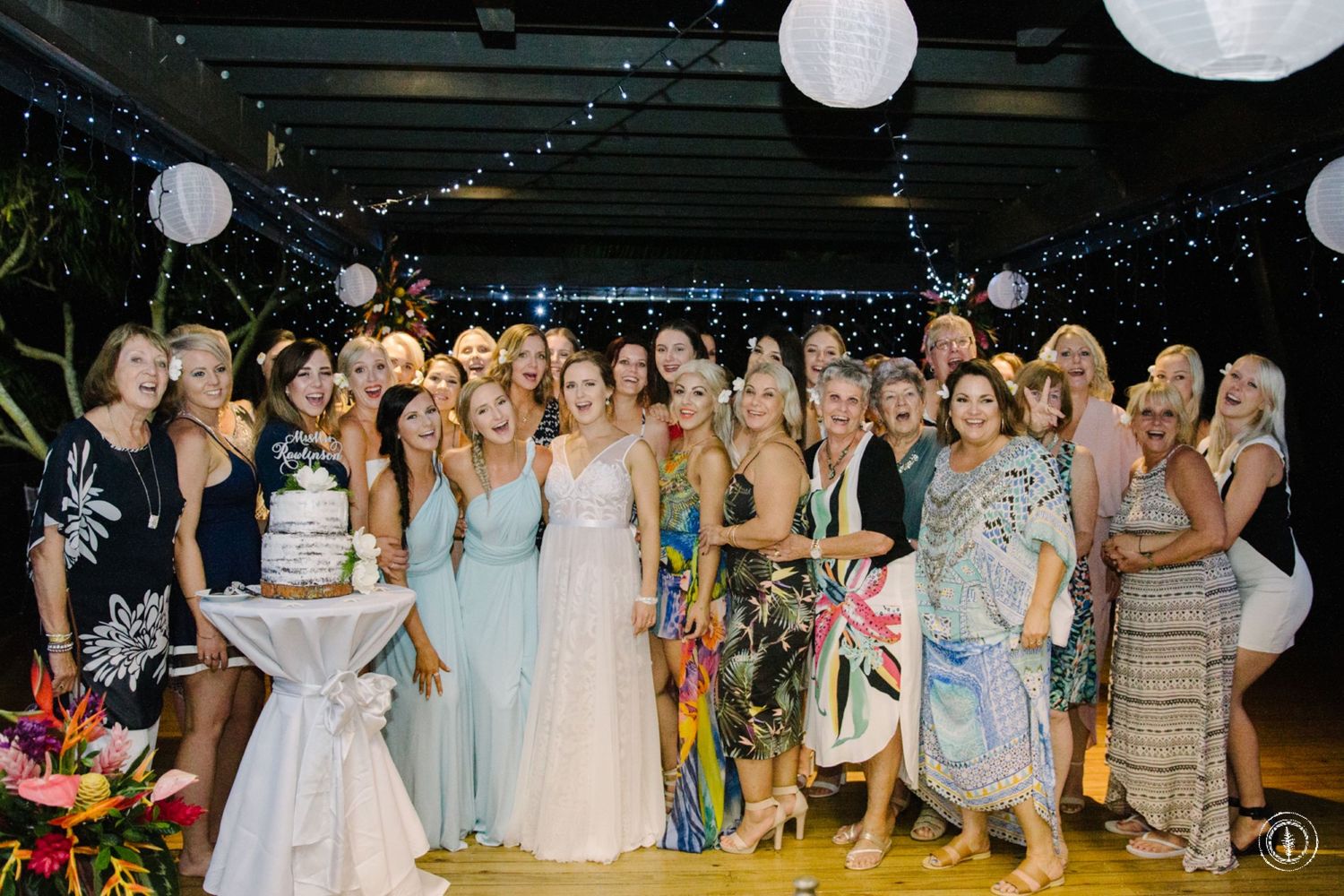 Large group photo of wedding attendees in formal attire with white paper lanterns and string lights overhead.