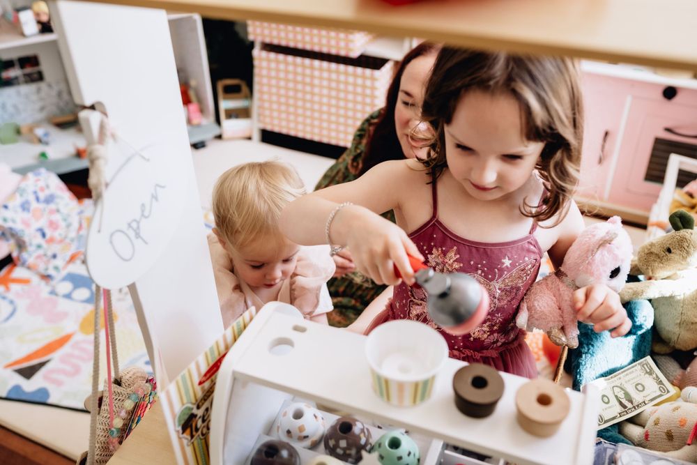 A girl playing with a toy kitchen with mum and young sister behind her