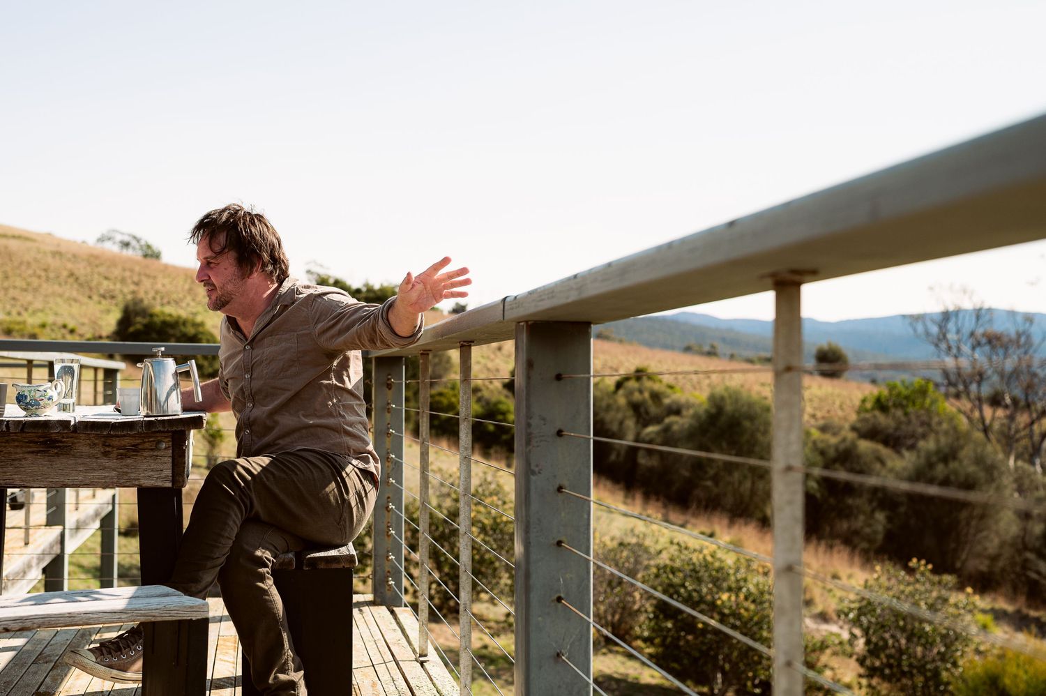 Person gesturing while standing on an outdoor deck with mountain views in the background.